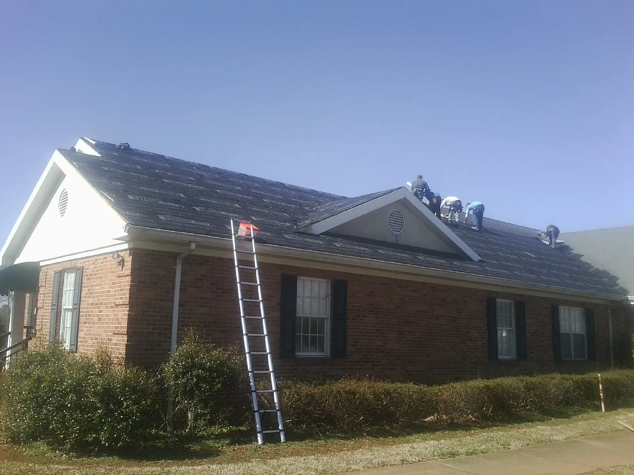 Crew tearing shingles off a brick ranch roof with a ladder set — Emergency Roof Repair work by our crew in West Mansfield