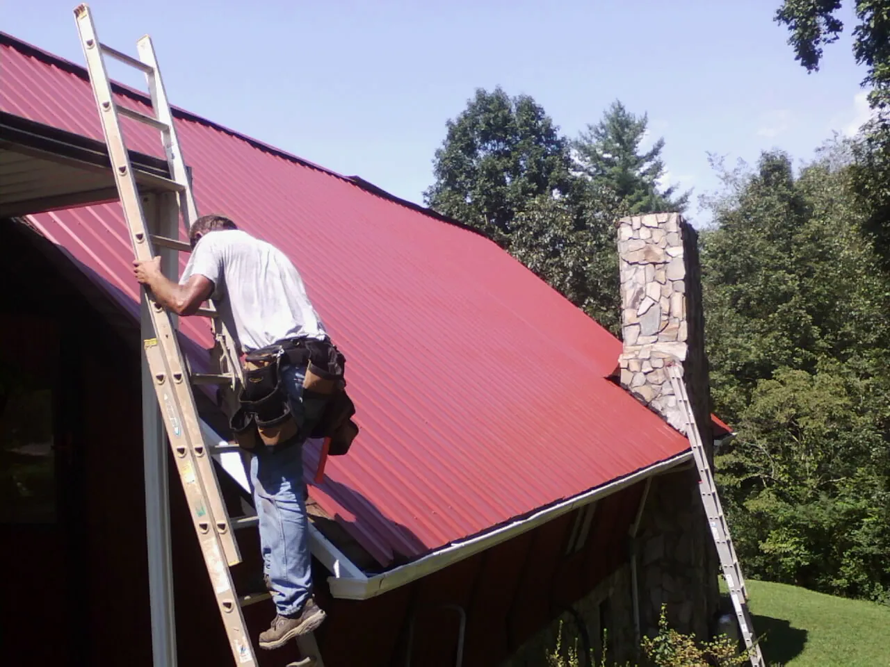 Roofer on a ladder beside a red metal roof and stone chimney — Soffit & Fascia Repair by Roofing Brothers in West Mansfield
